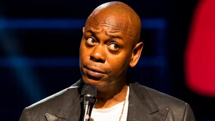 Dave Chappelle during his Netflix special looking questioningly off camera while wearing a black jacket and white shirt.